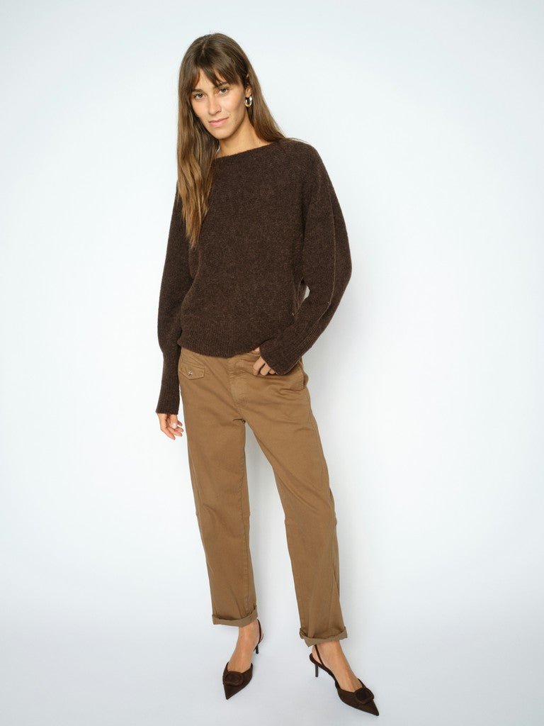 En kvinde i en mørkebrun sweater, MOS MOSH Aymen Camel-bukser og sorte højhælede sko, foran en ensfarvet lys baggrund.