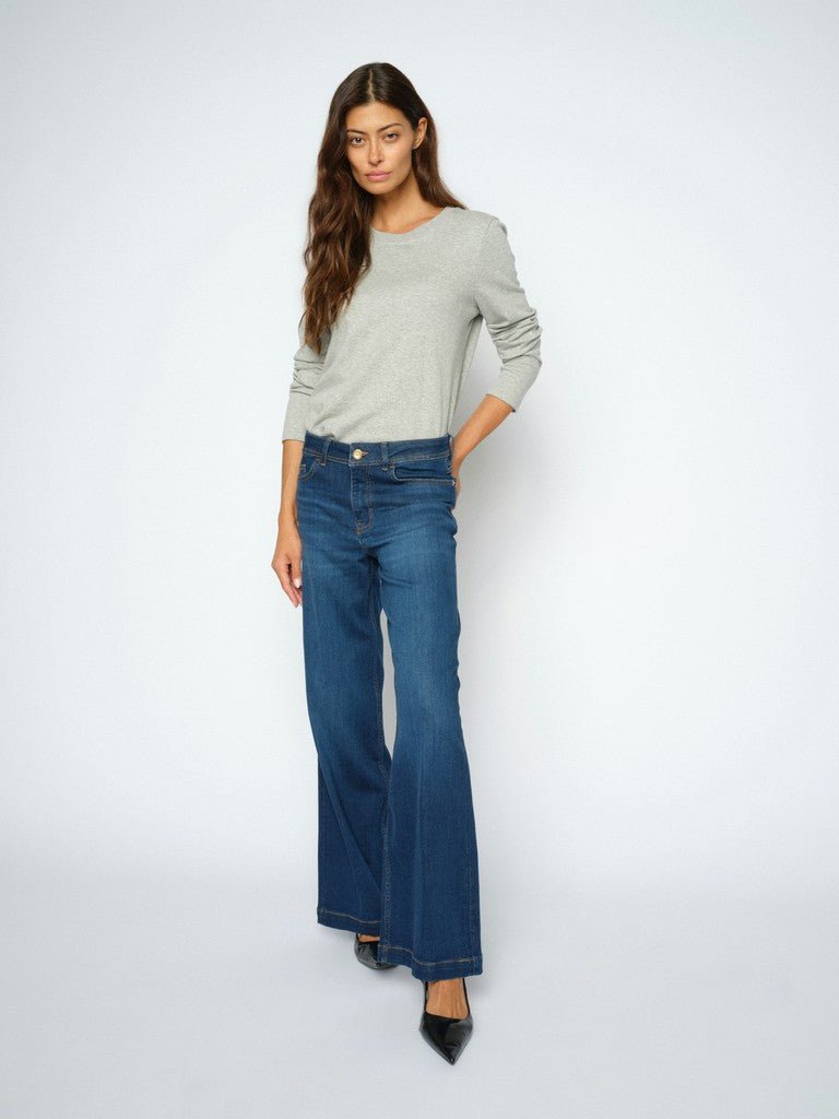 En kvinde iført en lysegrå top, MOS MOSH Dara Denim-jeans med brede ben og sorte højhælede sko står foran en ensfarvet baggrund.
