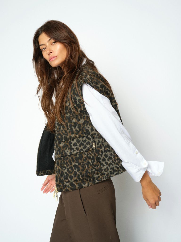 En kvinde iført en MOS MOSH Elise Leopard-vest, en hvid skjorte og brune bukser poserer foran en ensfarvet hvid baggrund.