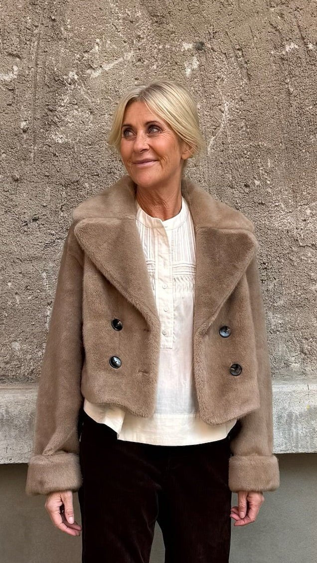 En ældre kvinde i en NOTYZ Nonice Lysebrun-jakke over en hvid bluse står ved en væg med struktur.