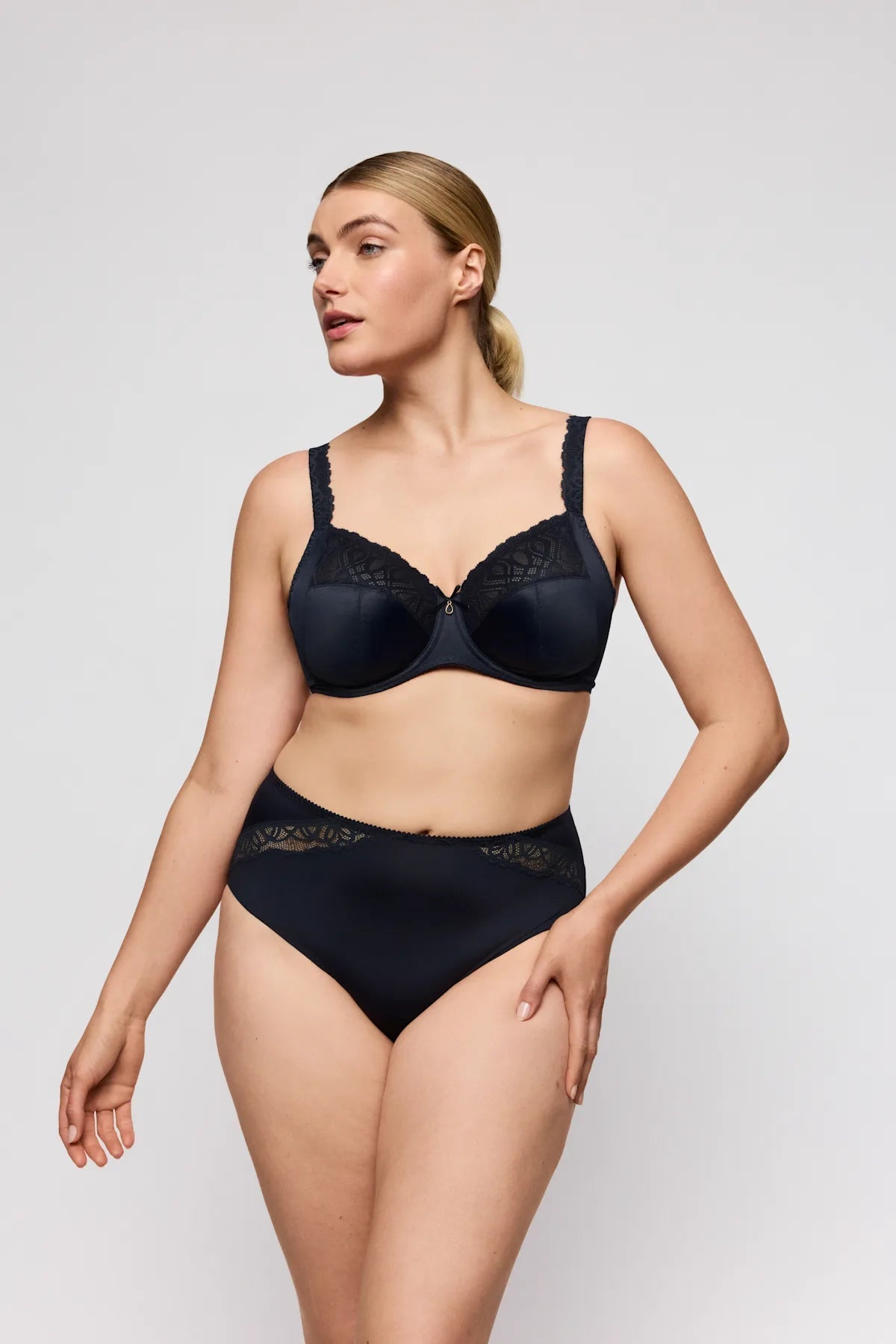 En kvinde iført et sæt bestående af en PrimaDonna Salerno-bh i farven Midnight Blue og et par højtaljede trusser står foran en ensfarvet lys baggrund.