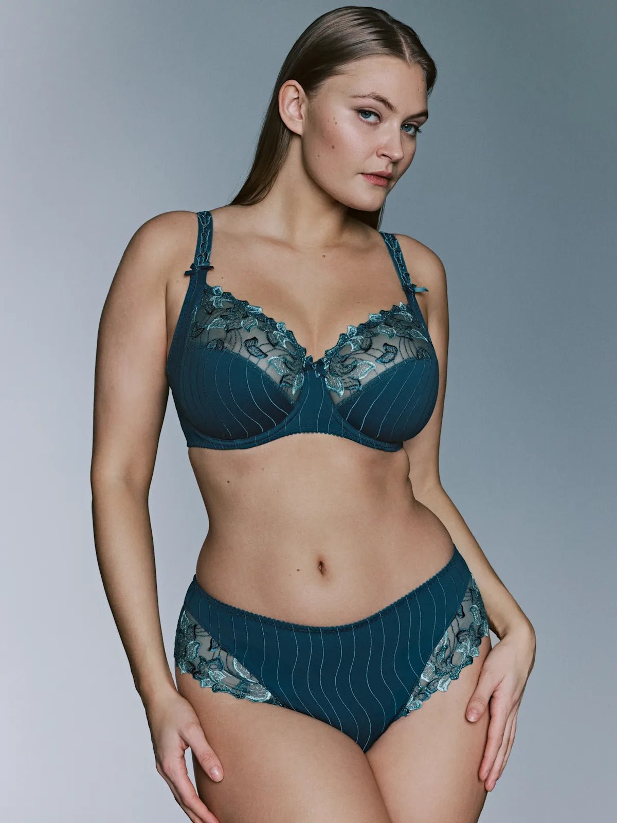 Model iført PrimaDonna - Deauville Peacock Petrol-sæt 155231, en bh med fulde skåle og bøjler, poserer foran en grå baggrund.