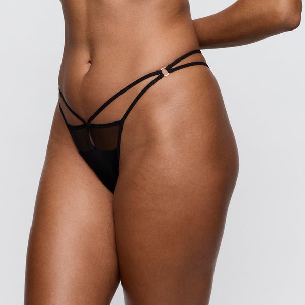 Model iført PrimaDonna Twist Sheary Black Sort-g-streng i gennemsigtigt net, fotograferet fra overkroppen til låret på lys baggrund.
