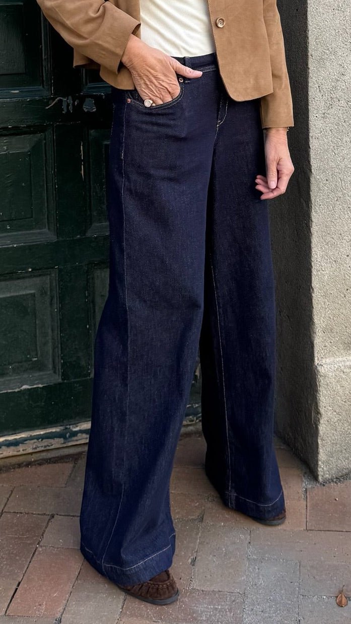 En person står på et fortov af mursten iført et par Red Button Claudine L 31-jeans i mørk denim, en lysebrun jakke og brune sko.