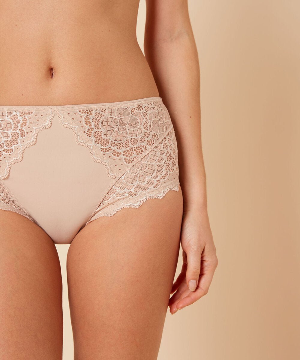 En kvinde har Simone Perele Caresse Peau Rosée Pudder-trusser på, i nudefarvet blonder, foran en beige baggrund, hvor overkroppen er synlig.