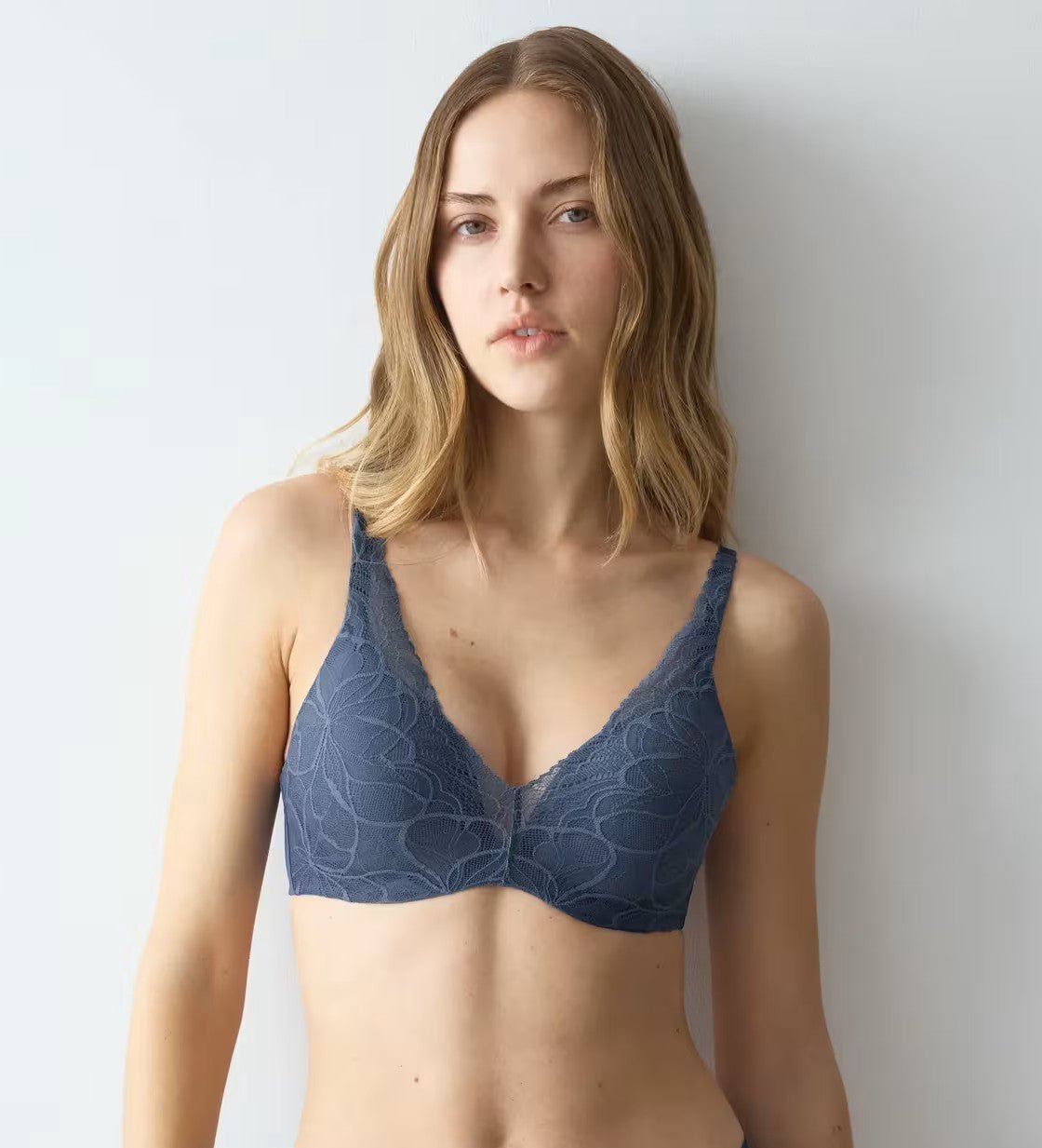 En kvinde iført en Triumph Body Make-Up Illusion Lace 787 Blå bh står foran en lys væg og ser selvsikkert ind i kameraet.
