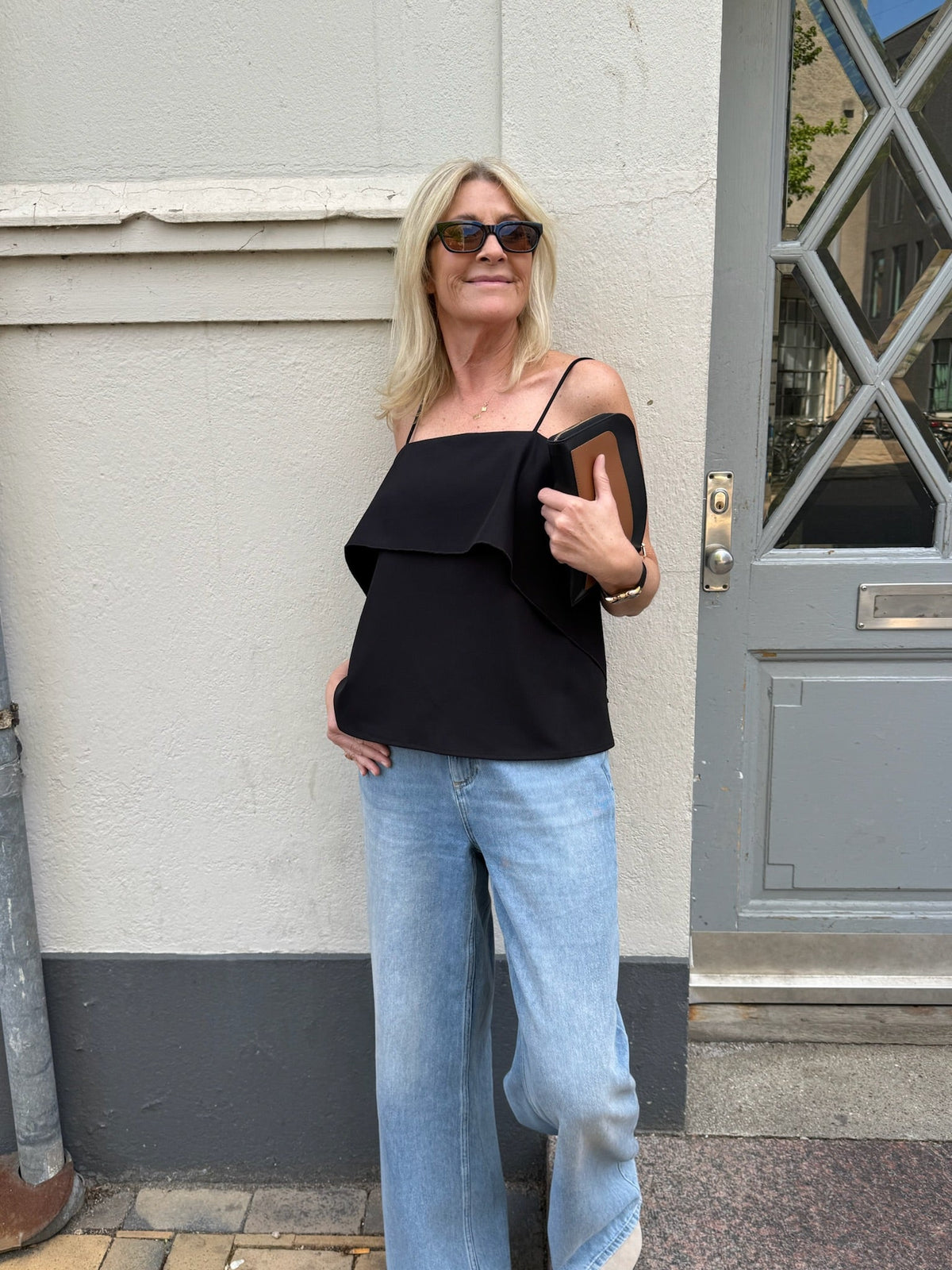 En smilende kvinde i solbriller og lyseblå jeans har en VILA Indira Sort-top på og står ved en grå dør med sin telefon i hånden.