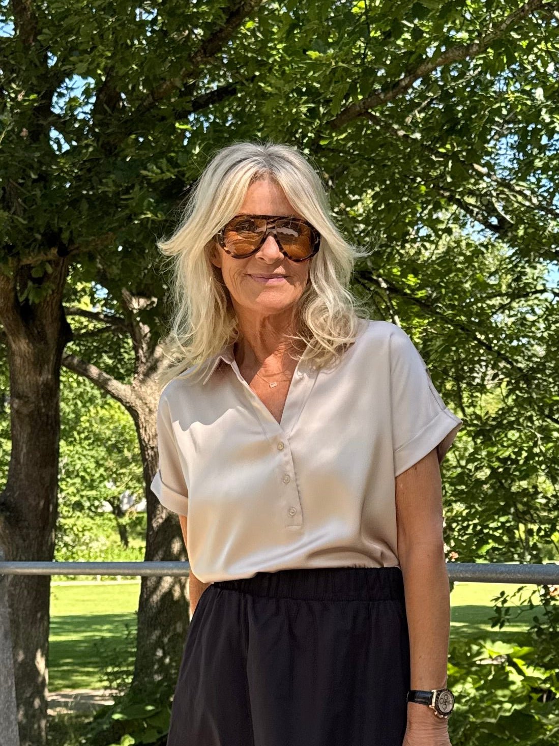 En kvinde med langt blondt hår har en VILA Vienna Ravenna Beige-bluse på og står ved nogle grønne træer på en solskinsdag.