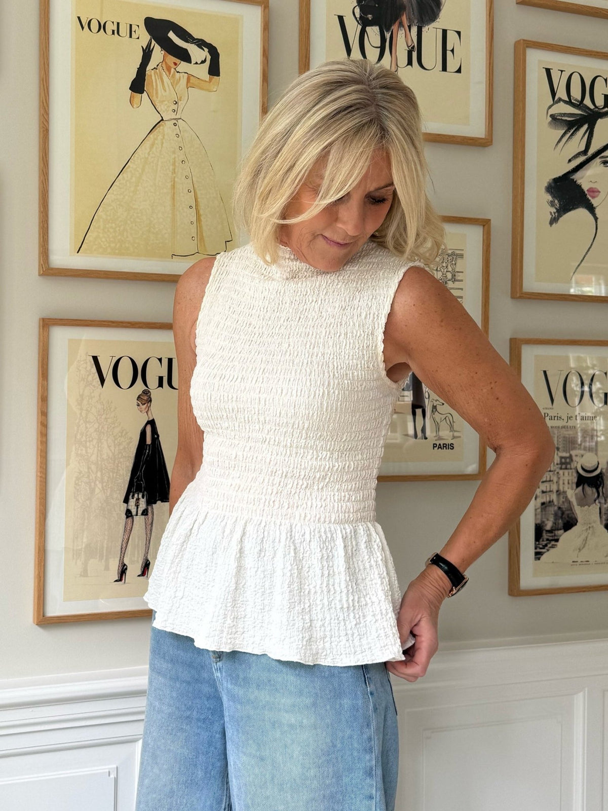 En kvinde iført en offwhite top og jeans fra VILA Sinne står foran indrammede forsidebilleder fra Vogue-magasinet på væggen.