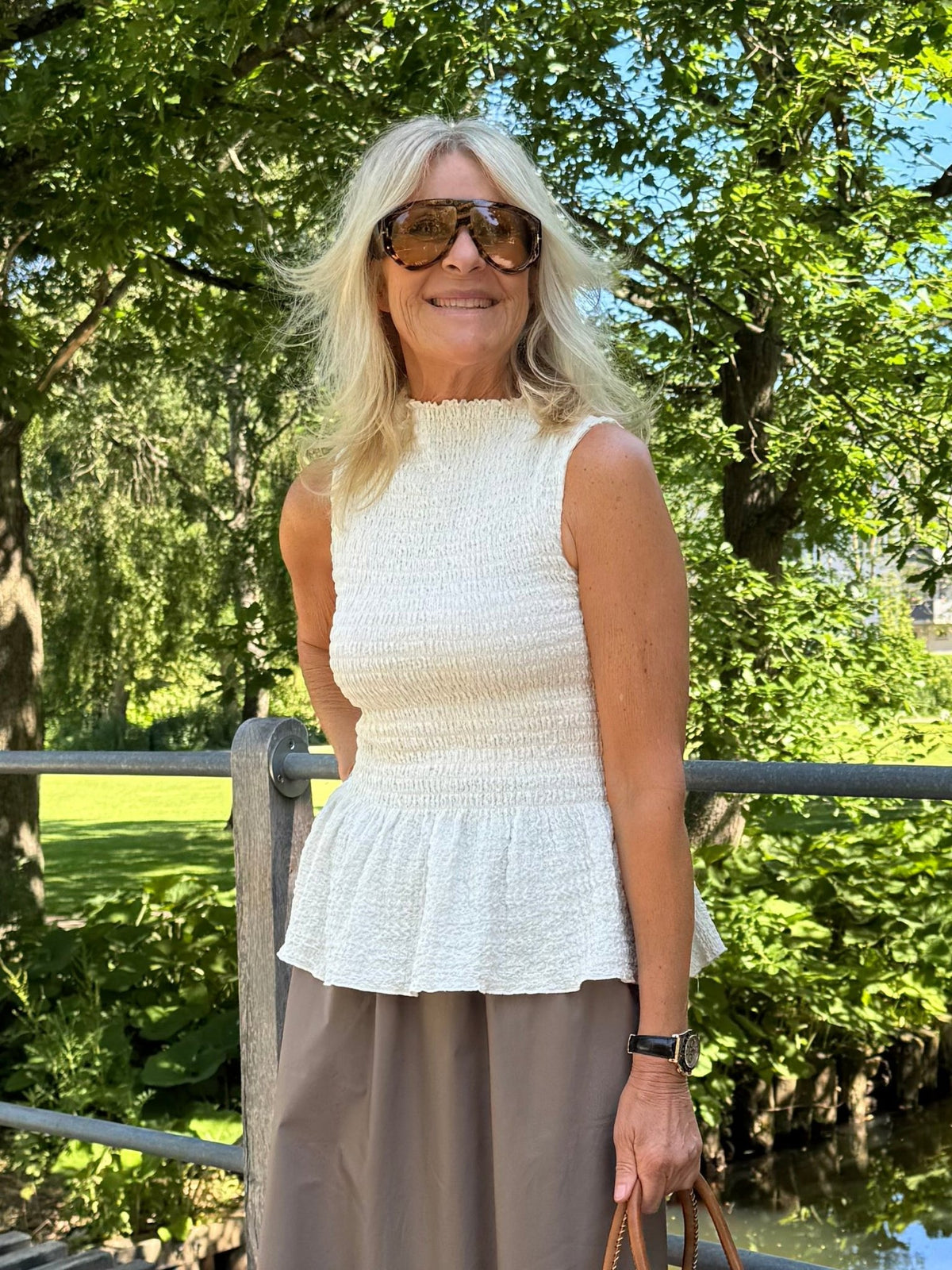 En blond kvinde iført en offwhite top og en grå nederdel fra VILA Sinne står udendørs ved et gelænder med solbriller på.