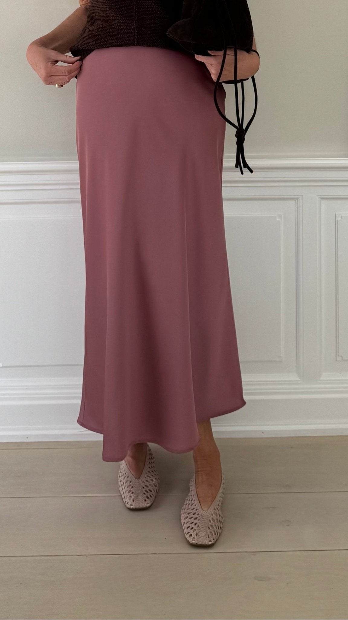 En person står på et lyst trægulv ved en hvid væg iført en brun bluse, en VILA Vieliette Rosa midi-nederdel og beige vævede sko.