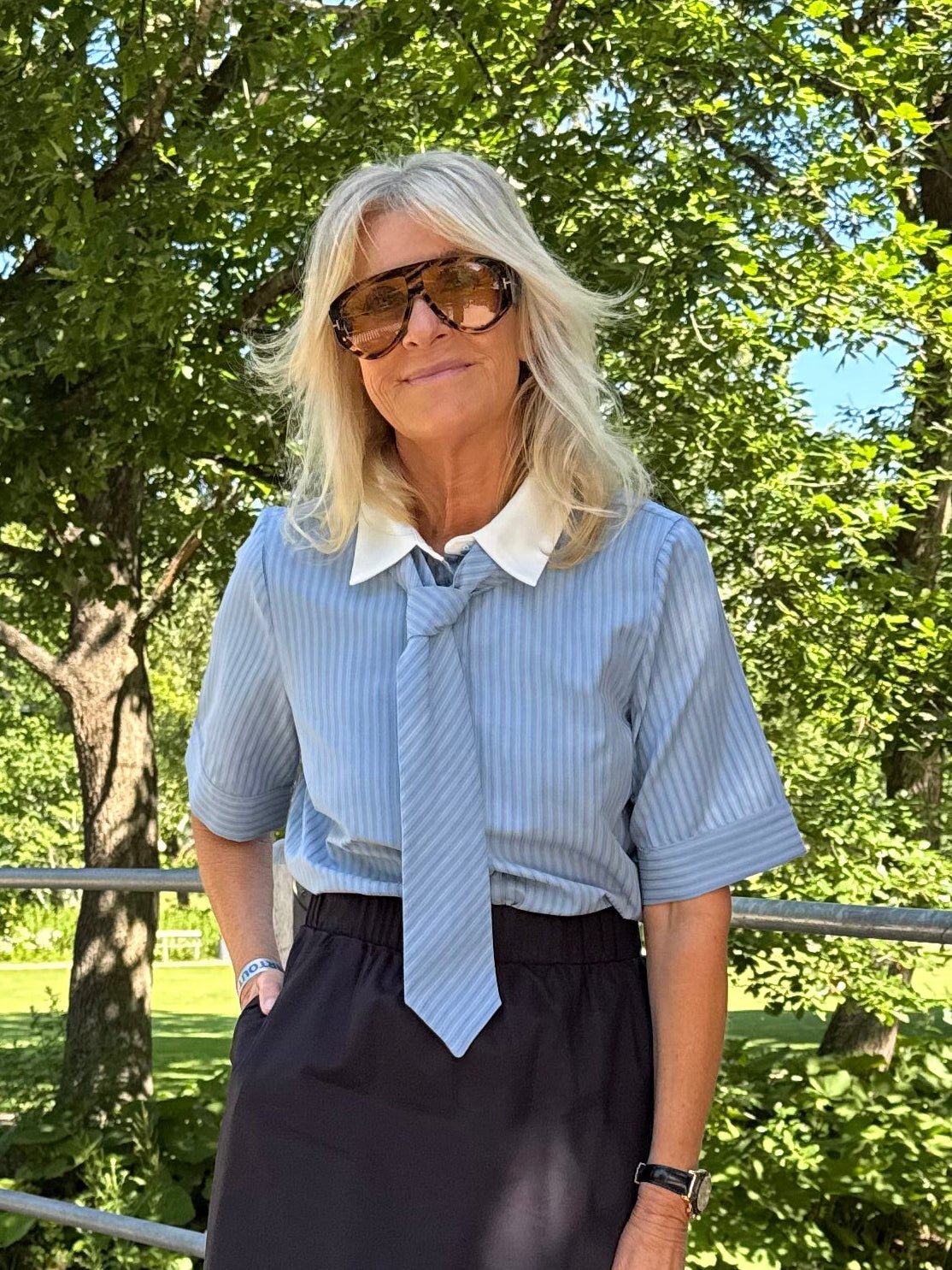 En blond kvinde med solbriller og en Linna Blå-bluse fra VILA står udendørs ved nogle træer iført en sort nederdel.