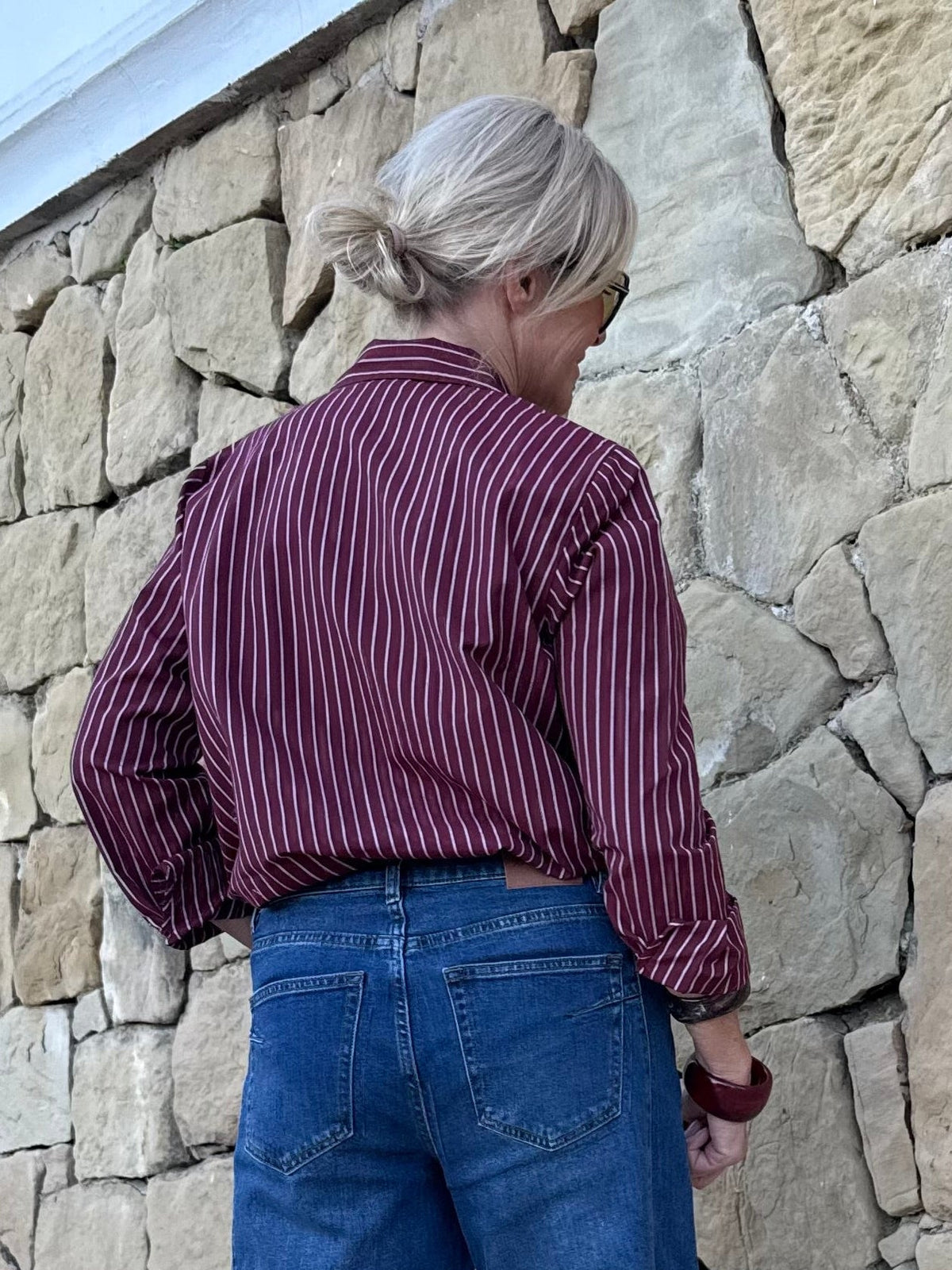 En person med blondt hår sat op i en knold, iført VILA Vidarma Bordeaux og blå jeans, står med ansigtet vendt mod en stenmur.