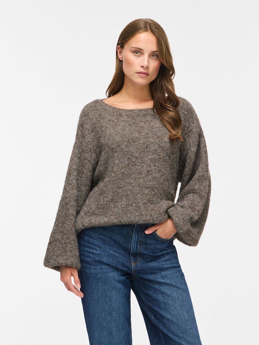 En kvinde i en løs VILA Vimarley Brunmeleret-top og blå jeans, der står foran en ensfarvet hvid baggrund.