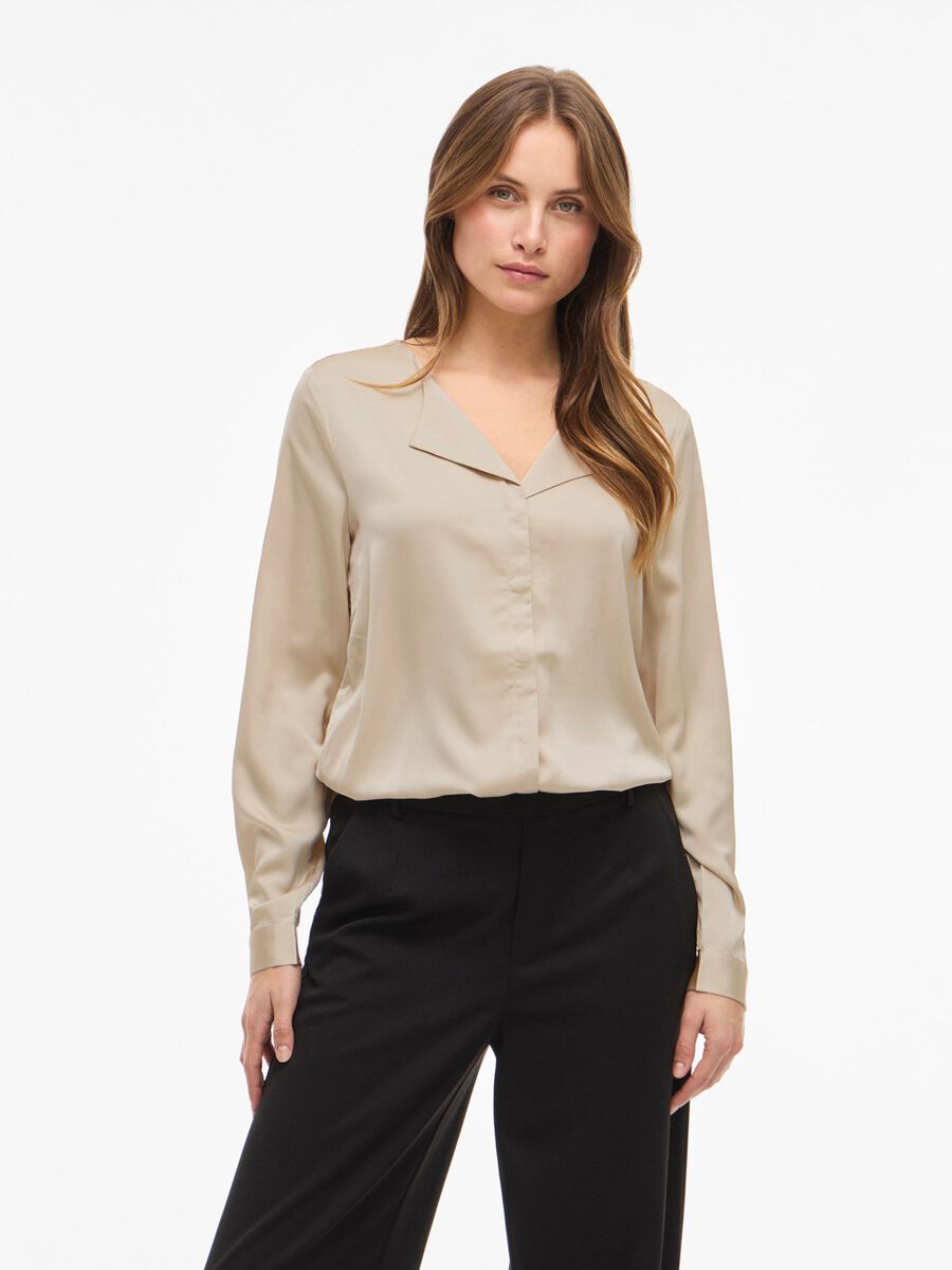 En kvinde iført en beige VILA Viellette 155327-bluse med V-udskæring og sorte bukser, der står foran en ensfarvet hvid baggrund.