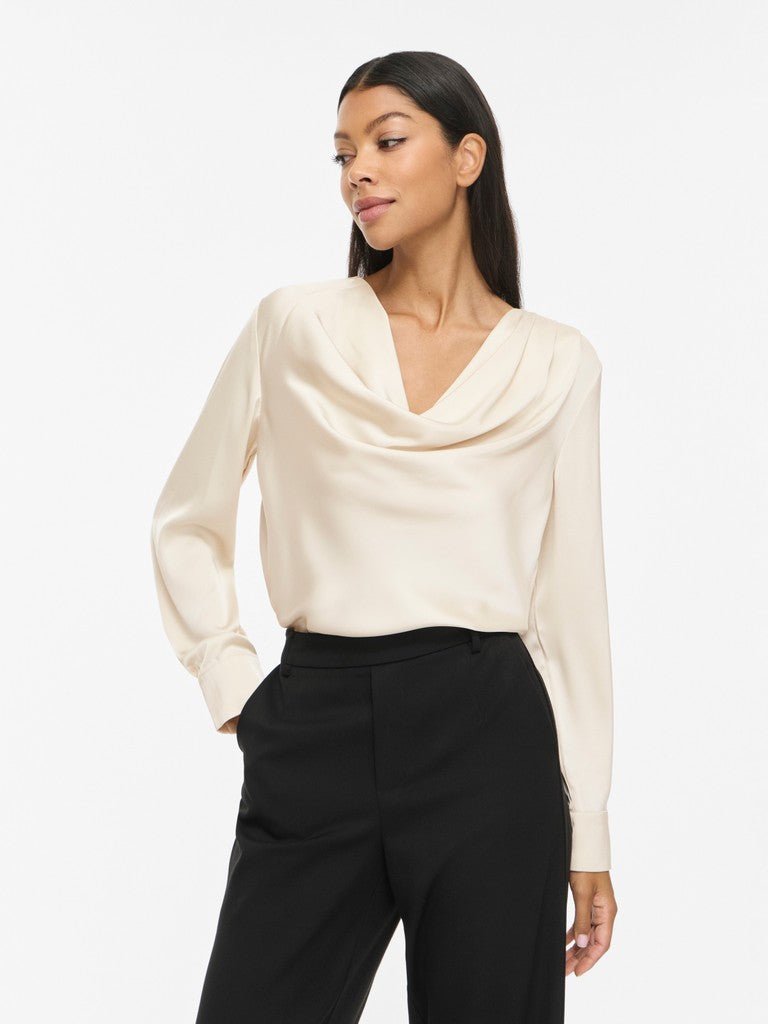 En kvinde iført en offwhite bluse og sorte bukser fra VILA VIFreja, der poserer med den ene hånd i lommen foran en ensfarvet baggrund.