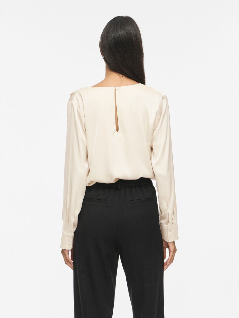 En kvinde står med ryggen til kameraet iført en offwhite bluse og sorte bukser fra VILA’s VIFreja-kollektion, foran en hvid baggrund.