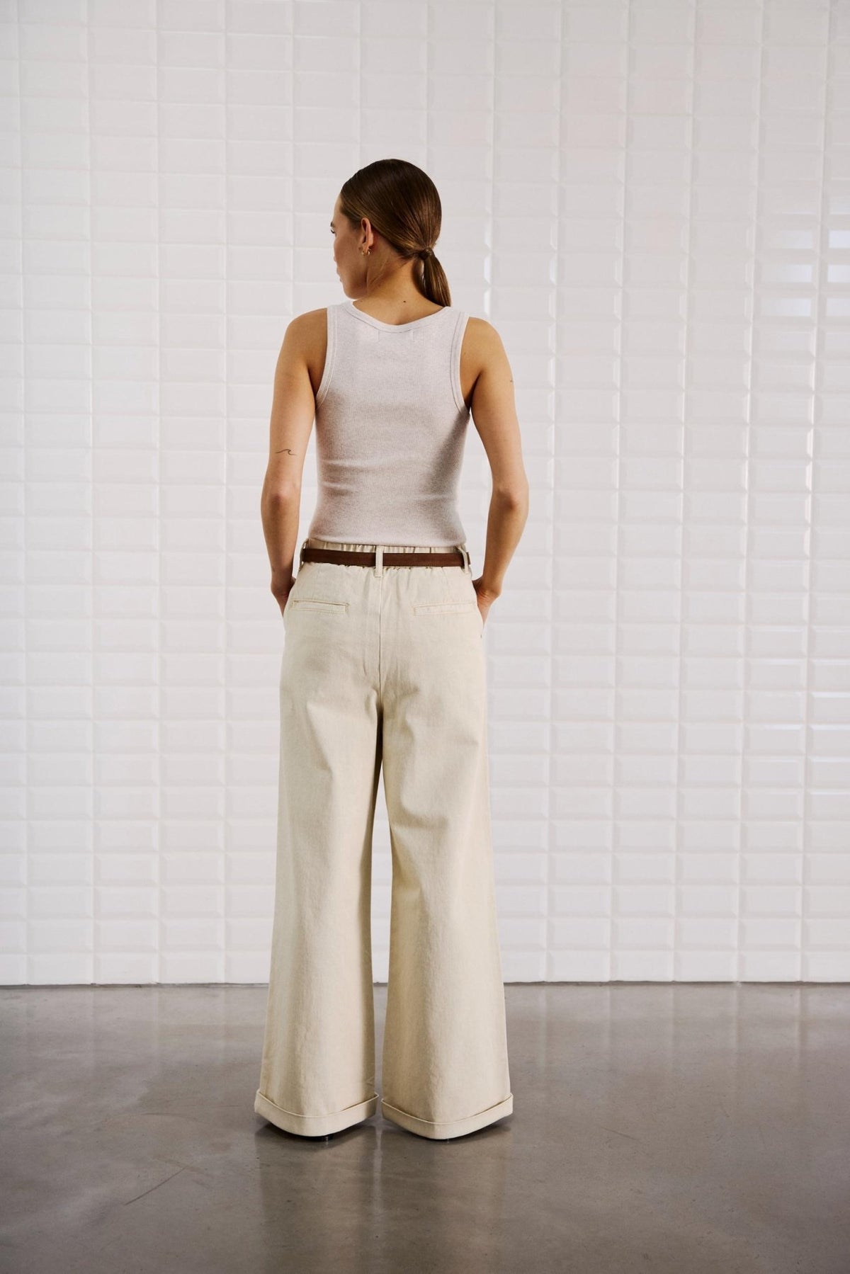En person, der står med ryggen til kameraet, iført en offwhite Steffi-tanktop og brede jeans fra WithBlack, i et minimalistisk rum.