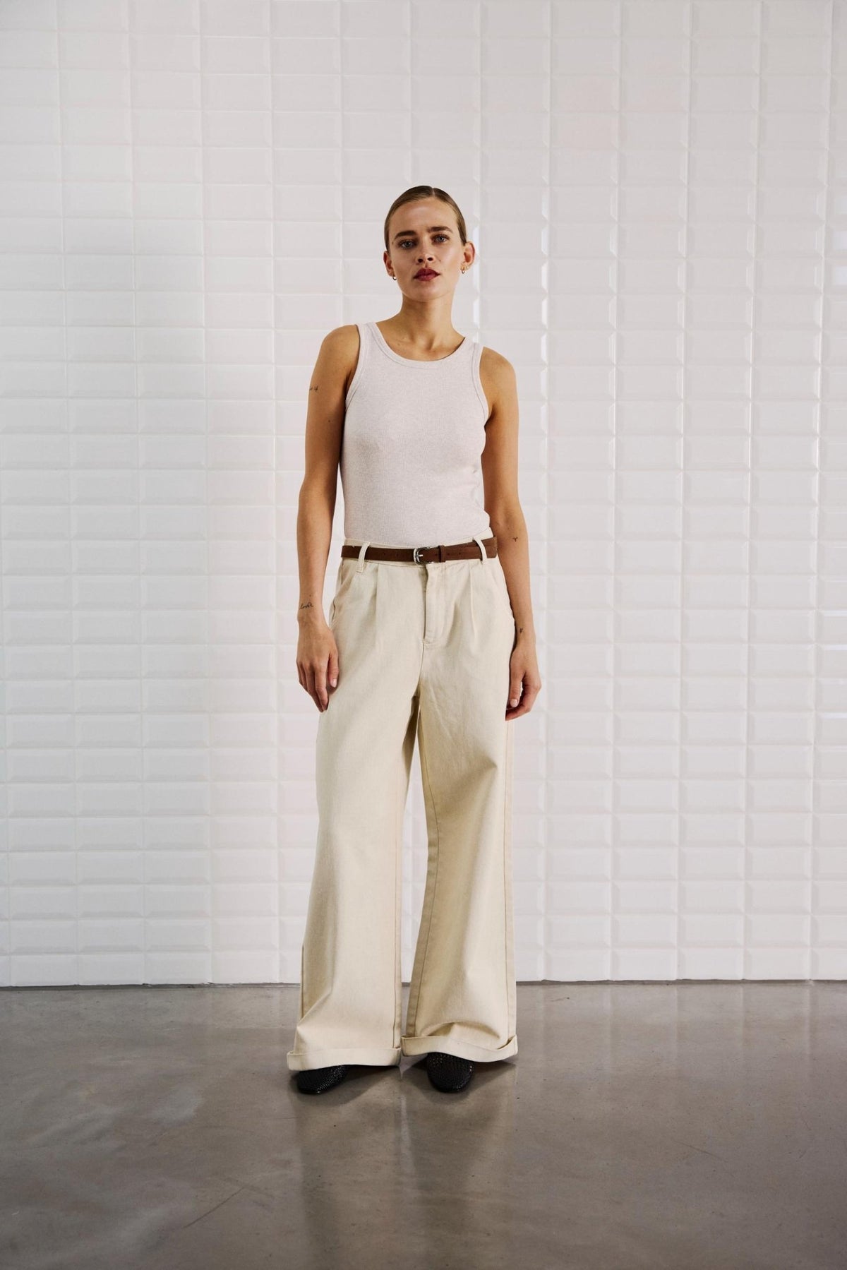 En person står foran en hvid flisebelagt væg iført WithBlack Steffi Off White-jeans, en hvid tanktop og sorte sko.