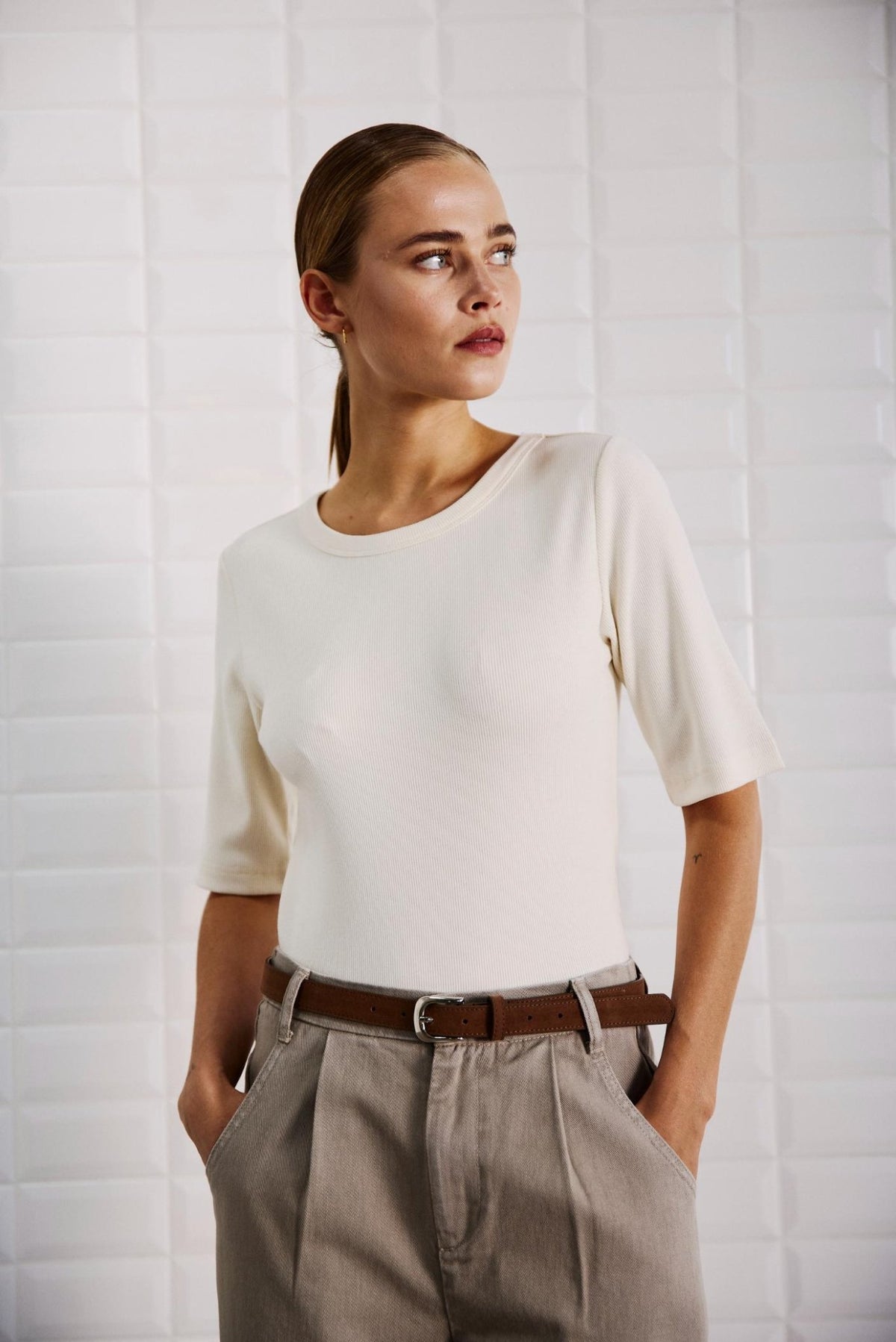 En kvinde iført en sort Lullu-T-shirt i offwhite med ribkant og beige bukser står ved en hvid flisebelagt væg med hænderne i lommerne.
