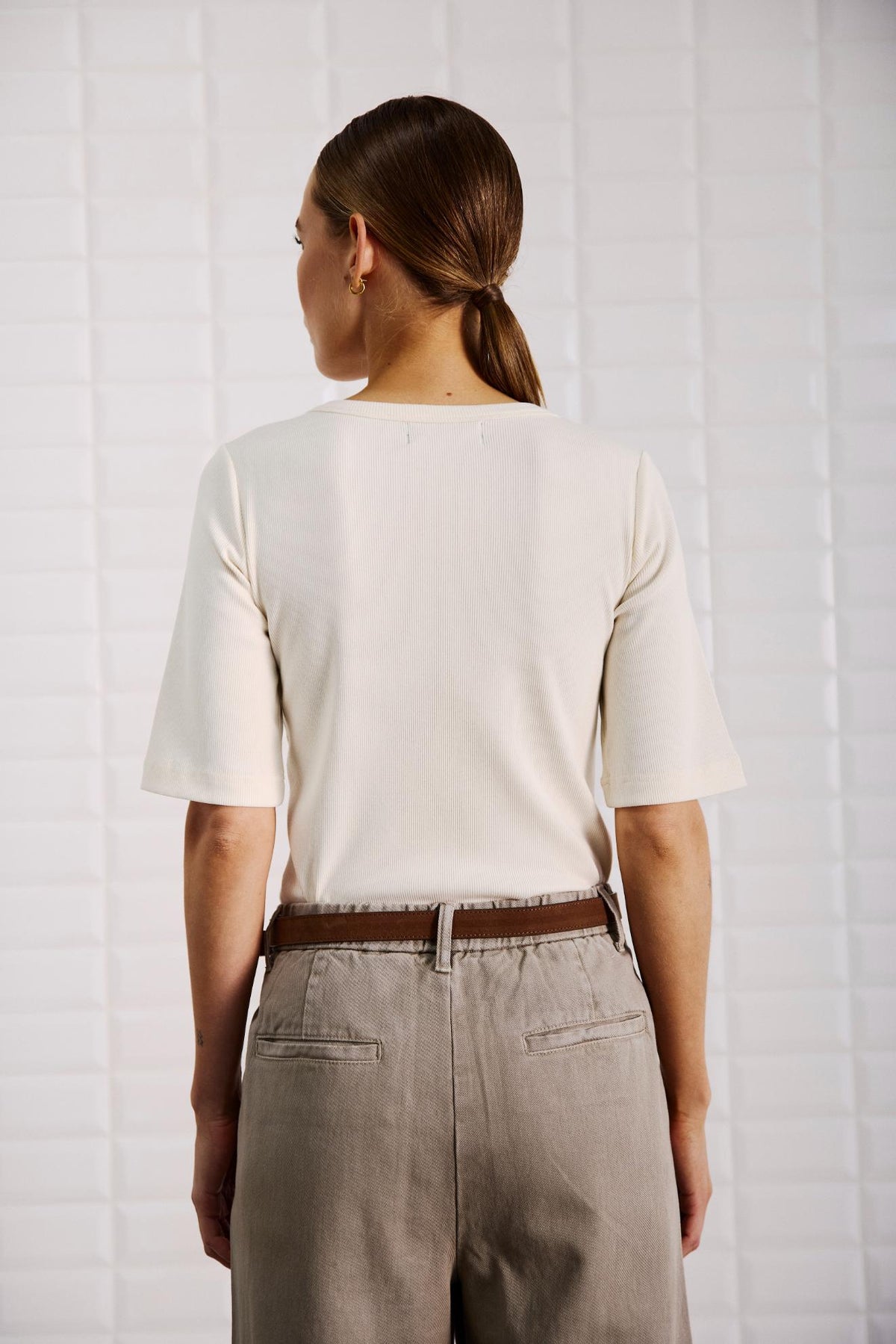 En person med brunt hår i en hestehale har en offwhite top fra WithBlack Lullu og beige bukser på og står med ryggen til kameraet.