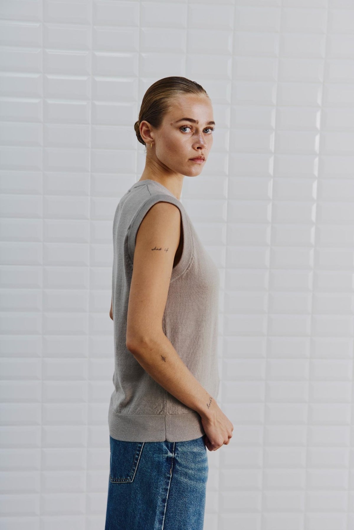 En kvinde iført en vest fra WithBlack – modellen »Miles Sand« – og blå jeans står med siden til ved en hvid flisebelagt væg og ser mod kameraet.