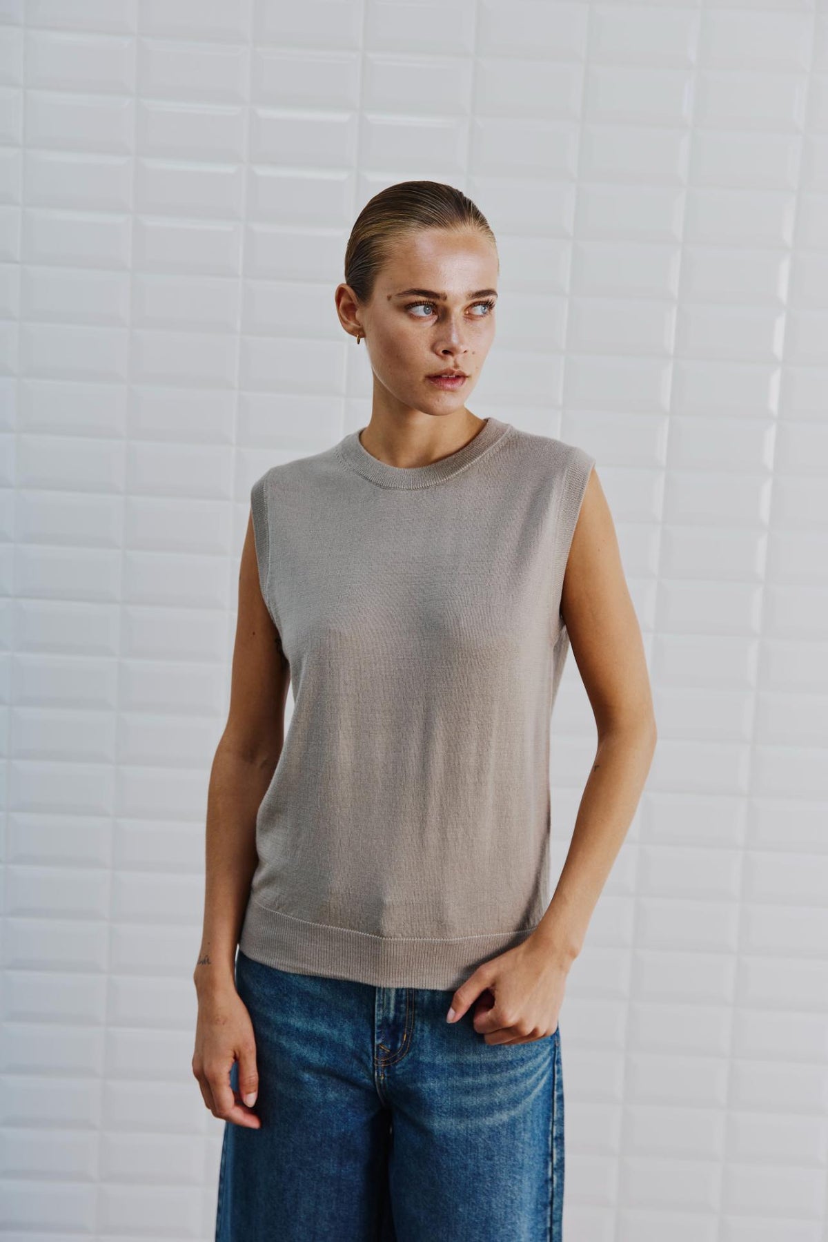 En person iført en WithBlack Miles Sand-vest og blå jeans står foran hvide fliser og kigger til siden.