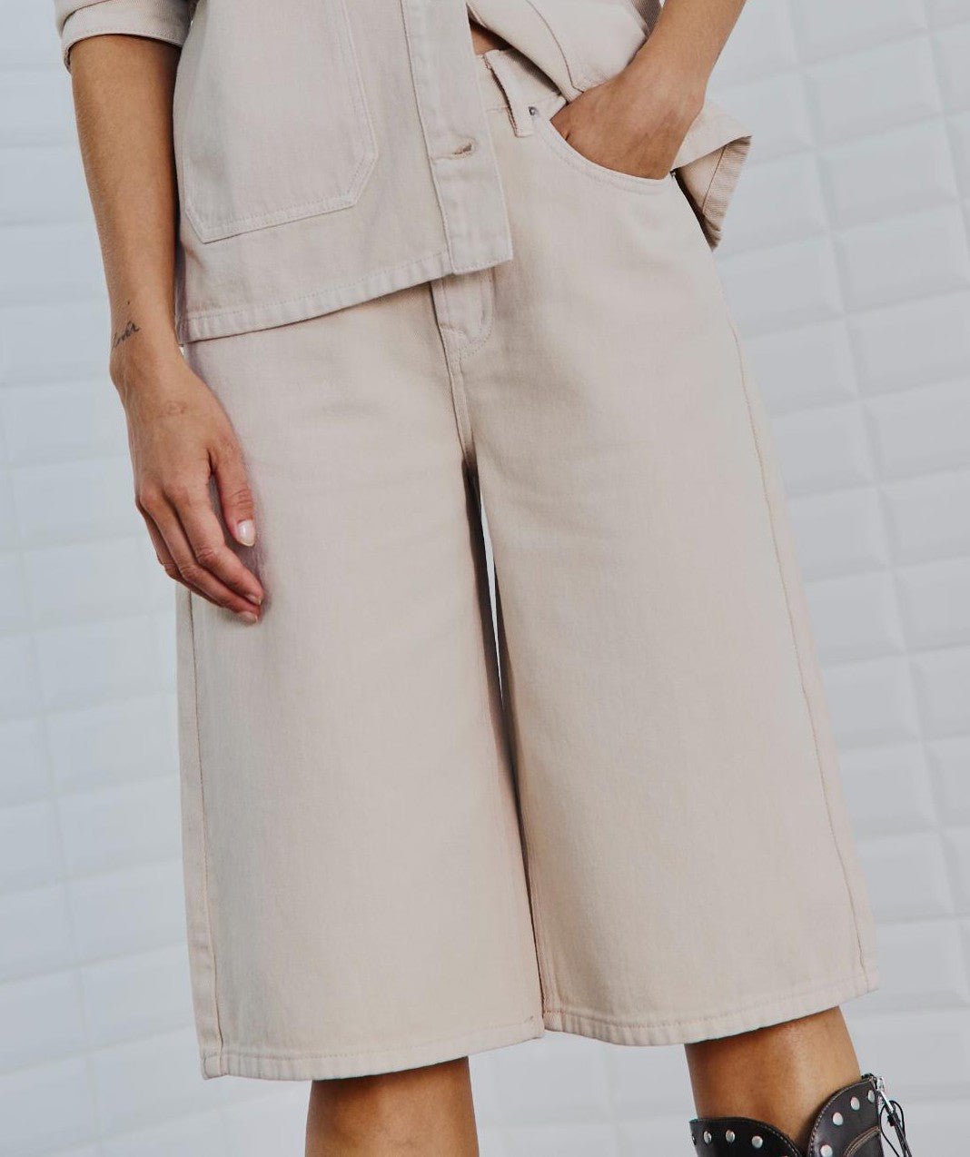 En model iført shorts og jakke i farven »Gala Beige« fra WithBlack, kombineret med sorte sandaler med nitter, står ved en flisebelagt væg.