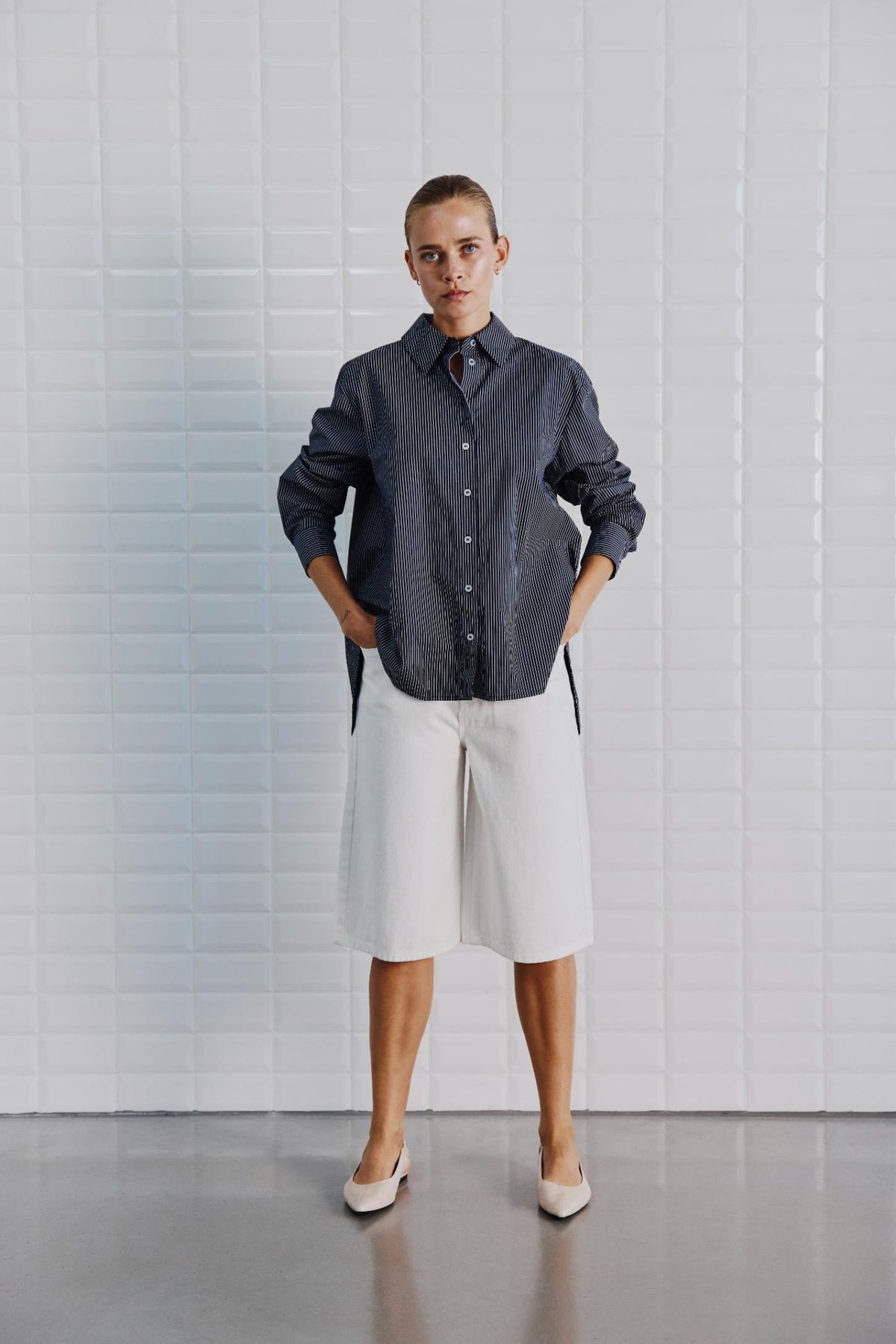 En person står ved en hvid flisebelagt væg iført en mørk stribet skjorte, Off White-shorts fra WithBlack Gala og beige sko.
