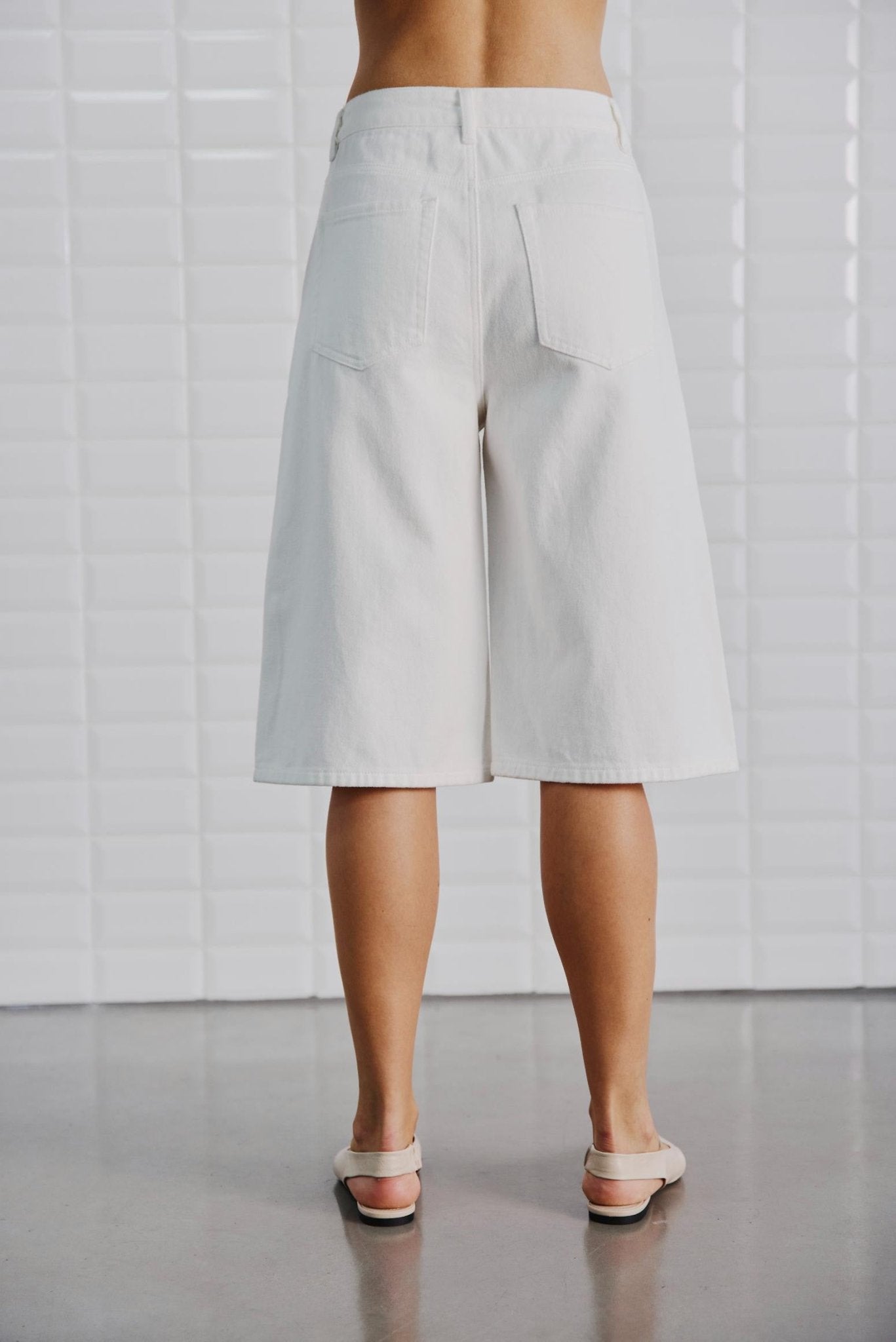 En person iført WithBlack – Gala-shorts i offwhite og sandaler står på et glat gulv med en flisebelagt væg bag sig.