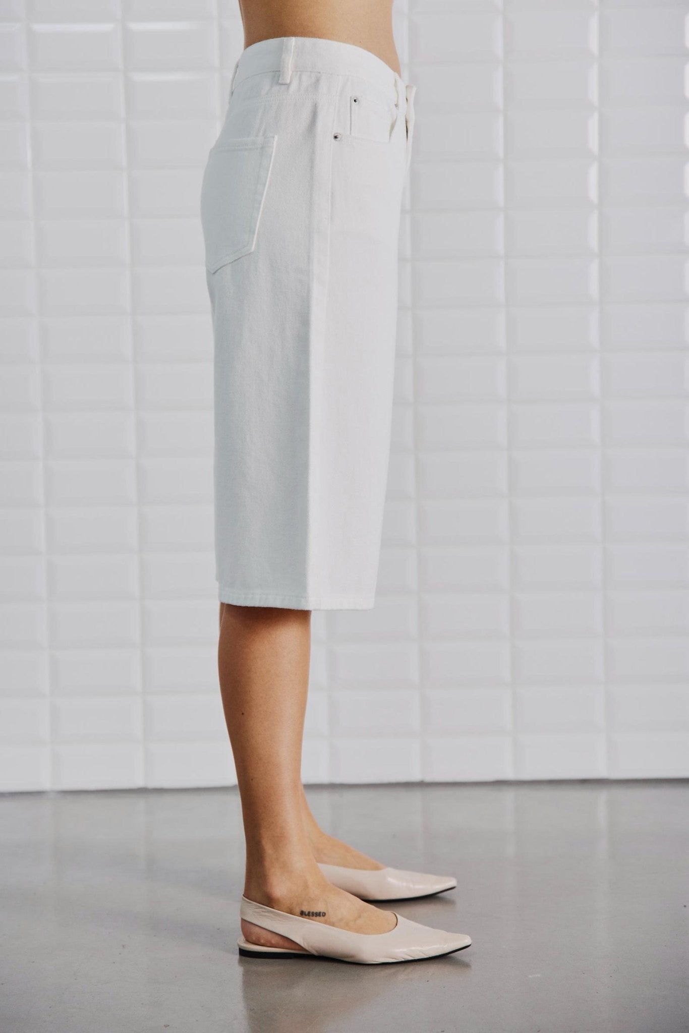 En person iført en Off-White-nederdel fra WithBlack og beige flade sko, der står med siden til en hvid flisebelagt væg.