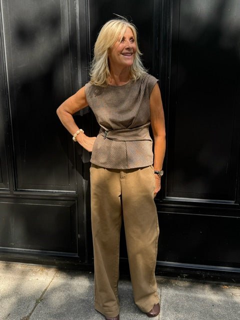 En blond kvinde i en brun bluse og WithBlack Steffi Camel-bukser står udenfor foran en sort mur.
