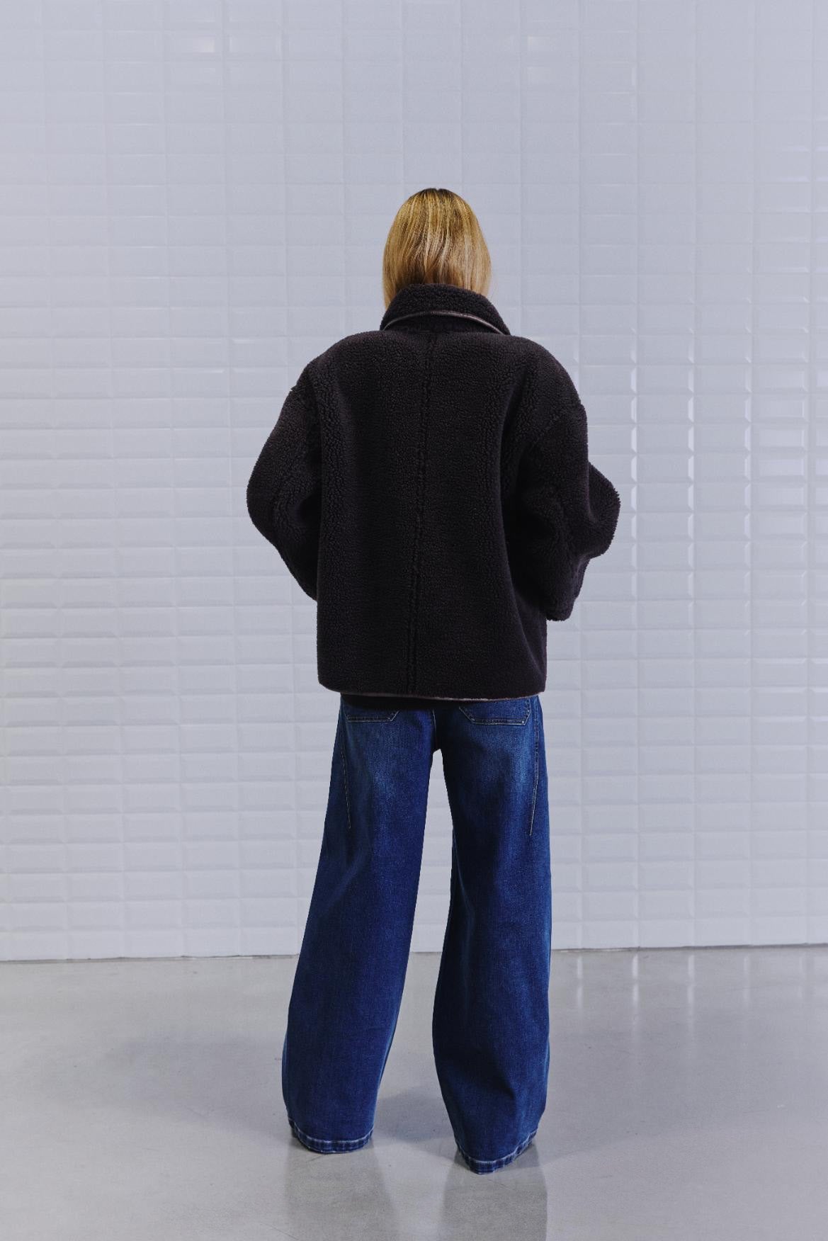 En person med skulderlangt hår iført en WithBlack IRIS Mørkebrun-jakke står med ansigtet vendt mod en hvid flisebelagt væg og har blå jeans med brede ben på.