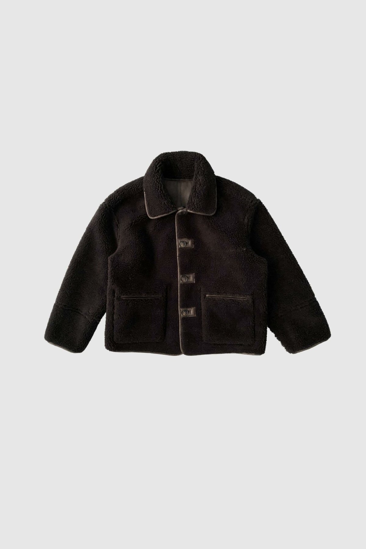 En sort IRIS-jakke i mørkebrun shearling med store knapper, lommer og krave, vist på en lys baggrund.