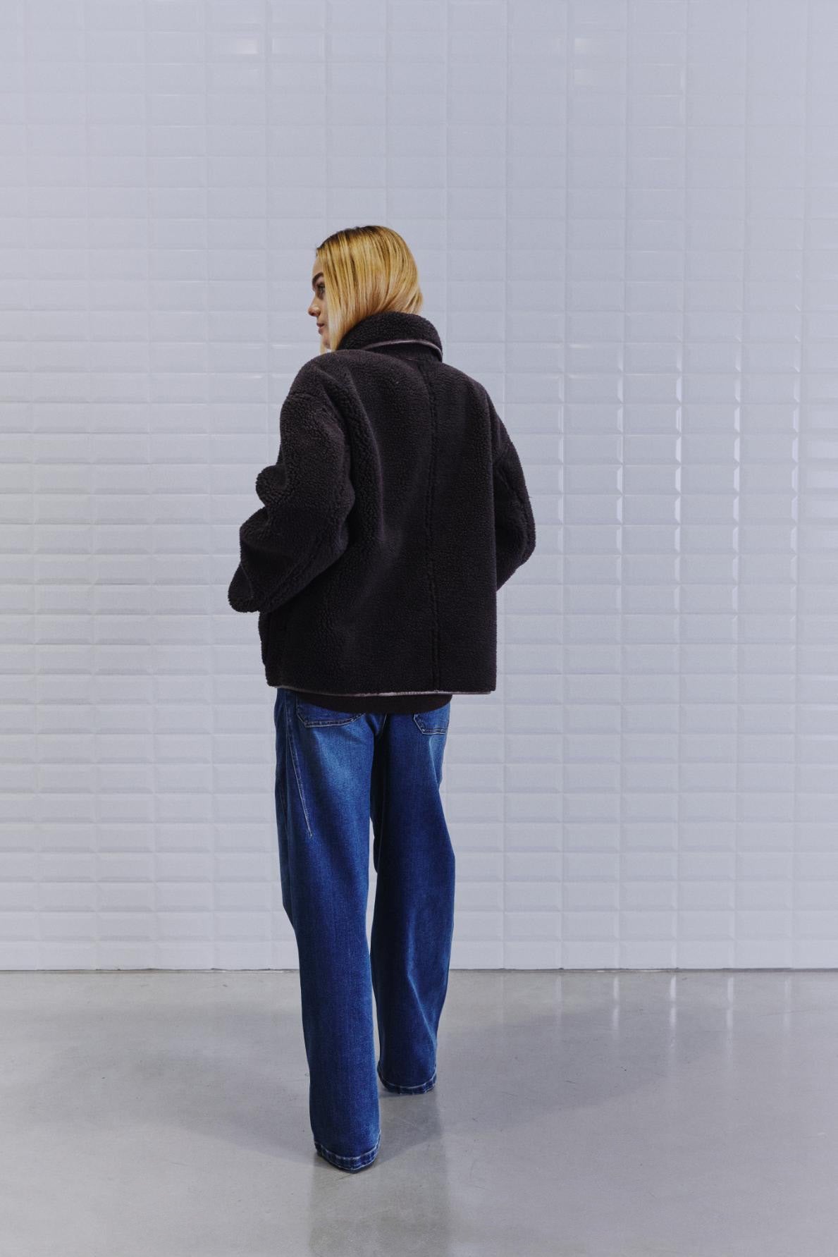 En person med blondt hår har en WithBlack IRIS Mørkebrun-jakke og blå jeans på og står ved en hvid flisebelagt væg.
