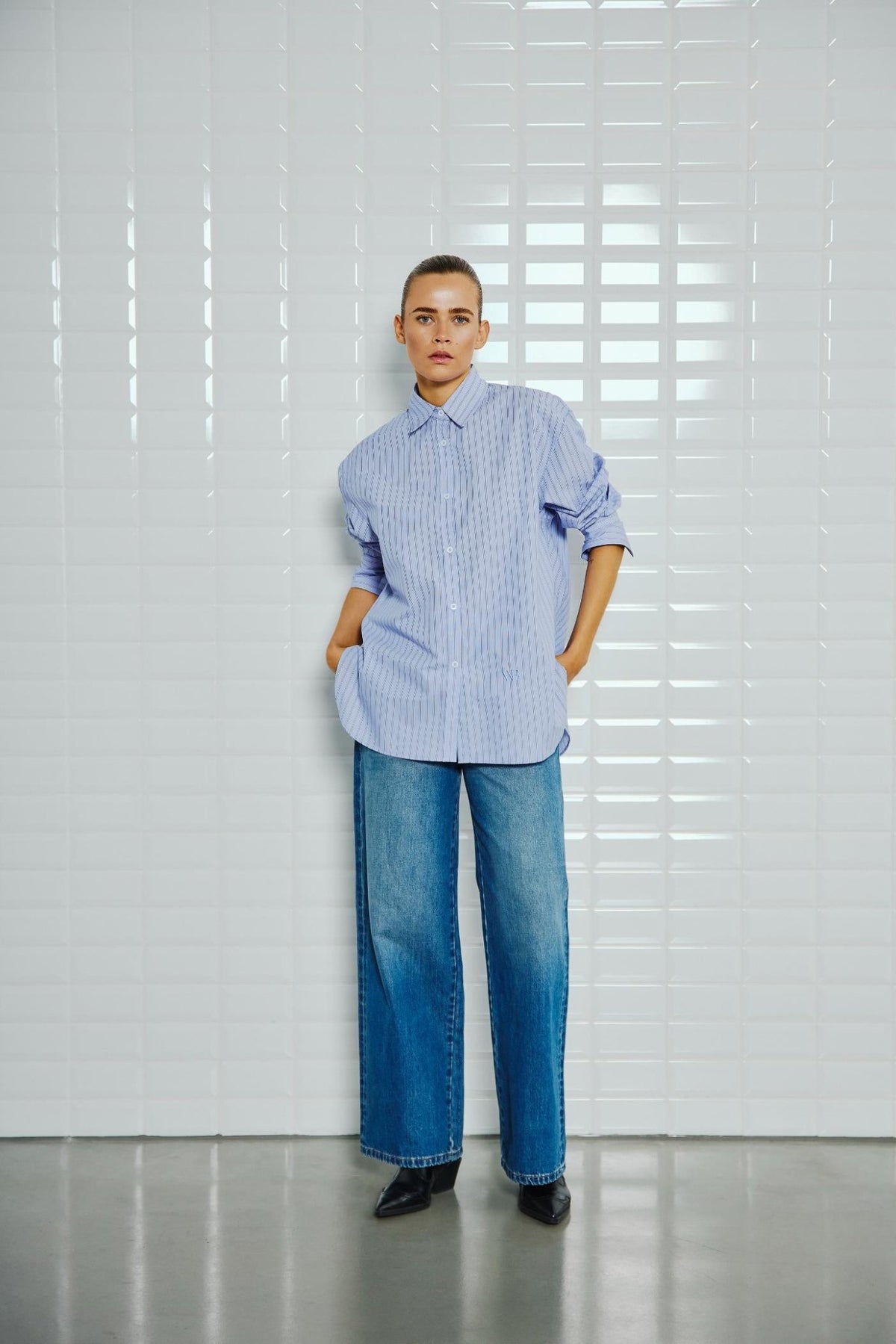 En person iført 156395 | WithBlack - Noa Blå, en blå skjorte med fine striber og knapper, blå jeans med brede ben og sorte støvler med spids snude.