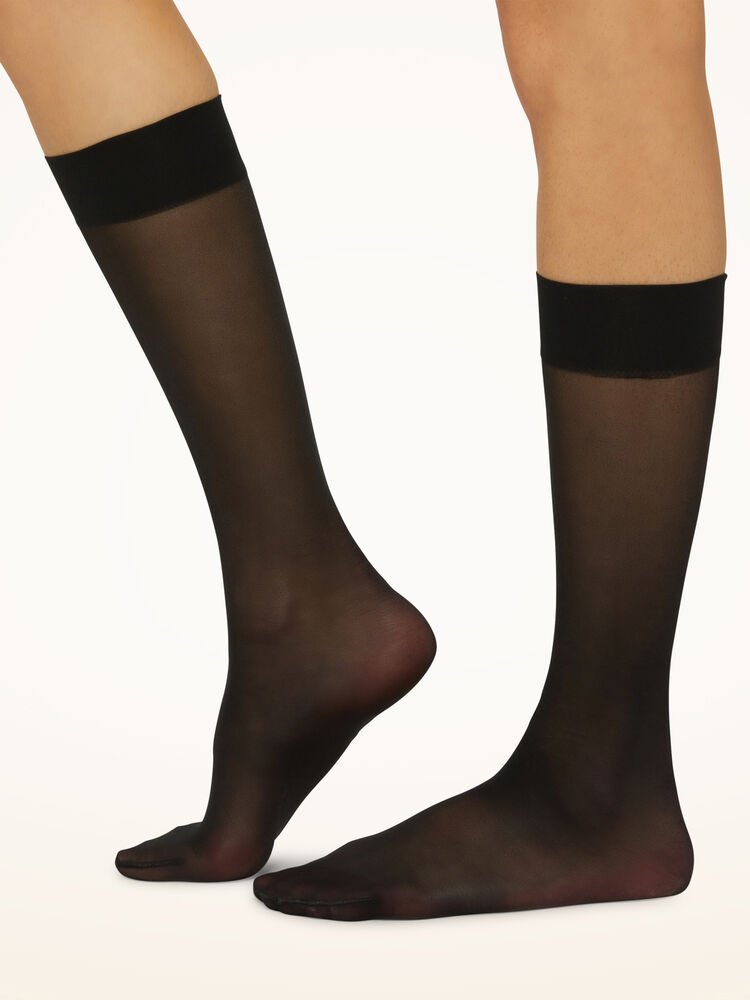 Fødder i Wolford Satin Touch 20 Black W knæstrømper med ensfarvede bånd står på en ensfarvet baggrund.