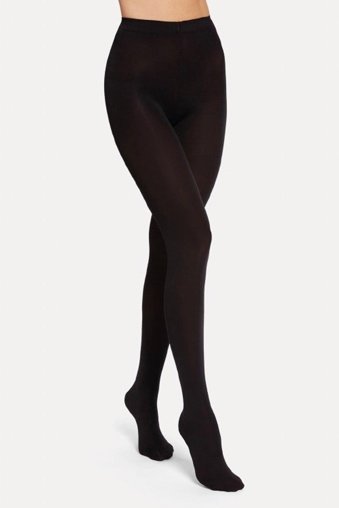 En model iført Wolford Opaque 70 Black W-strømpebukser står med det ene ben fremstrakt foran en ensfarvet lys baggrund.