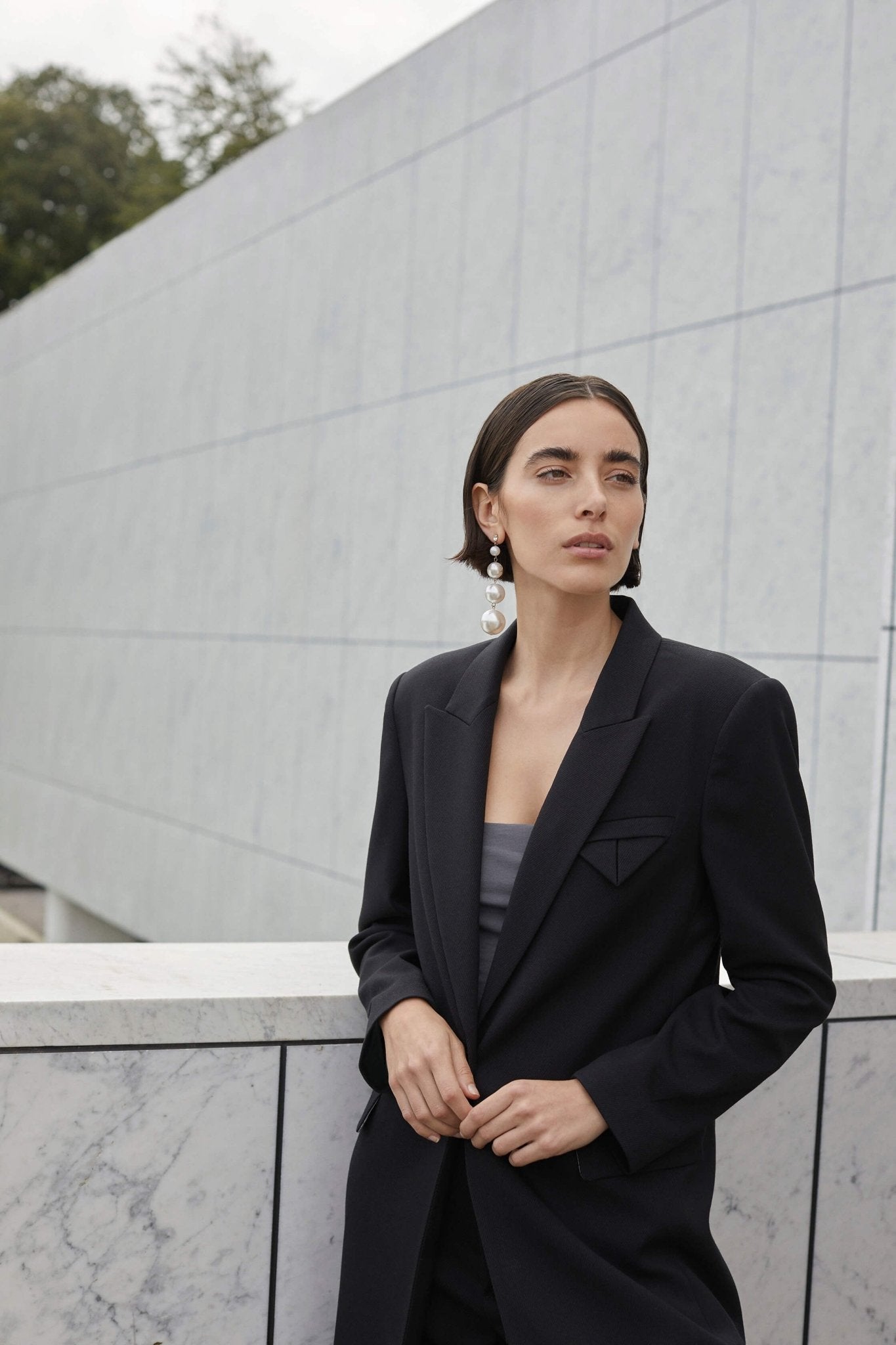 En kvinde har en sort blazer fra Copenhagen Muse Tailor på foran en marmorvæg med moderne arkitektur i baggrunden.
