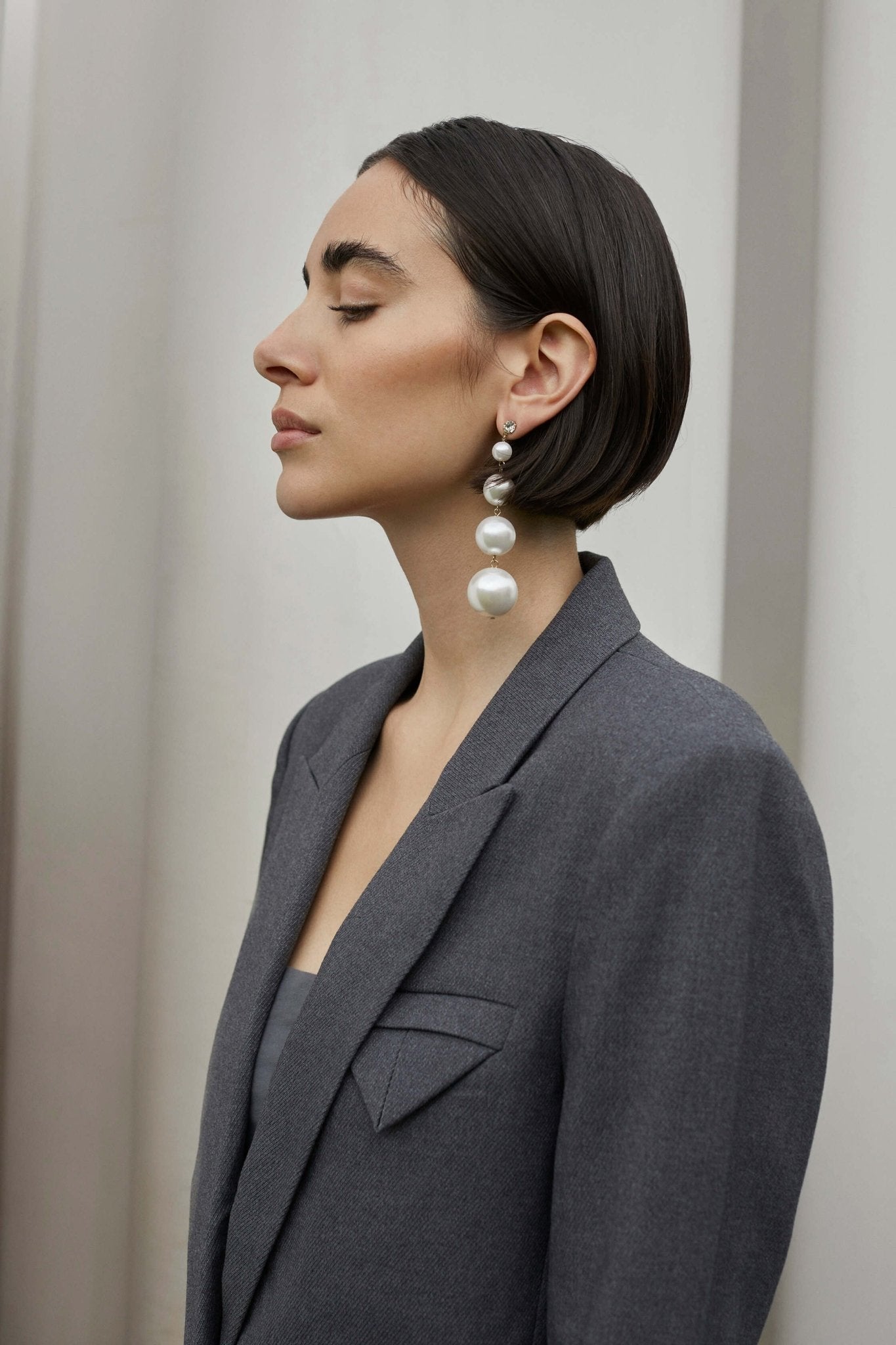 Kvinde iført en grå blazer fra Copenhagen Muse Tailor, kort mørkt hår, store perleøreringe, stående i profil med lukkede øjne.